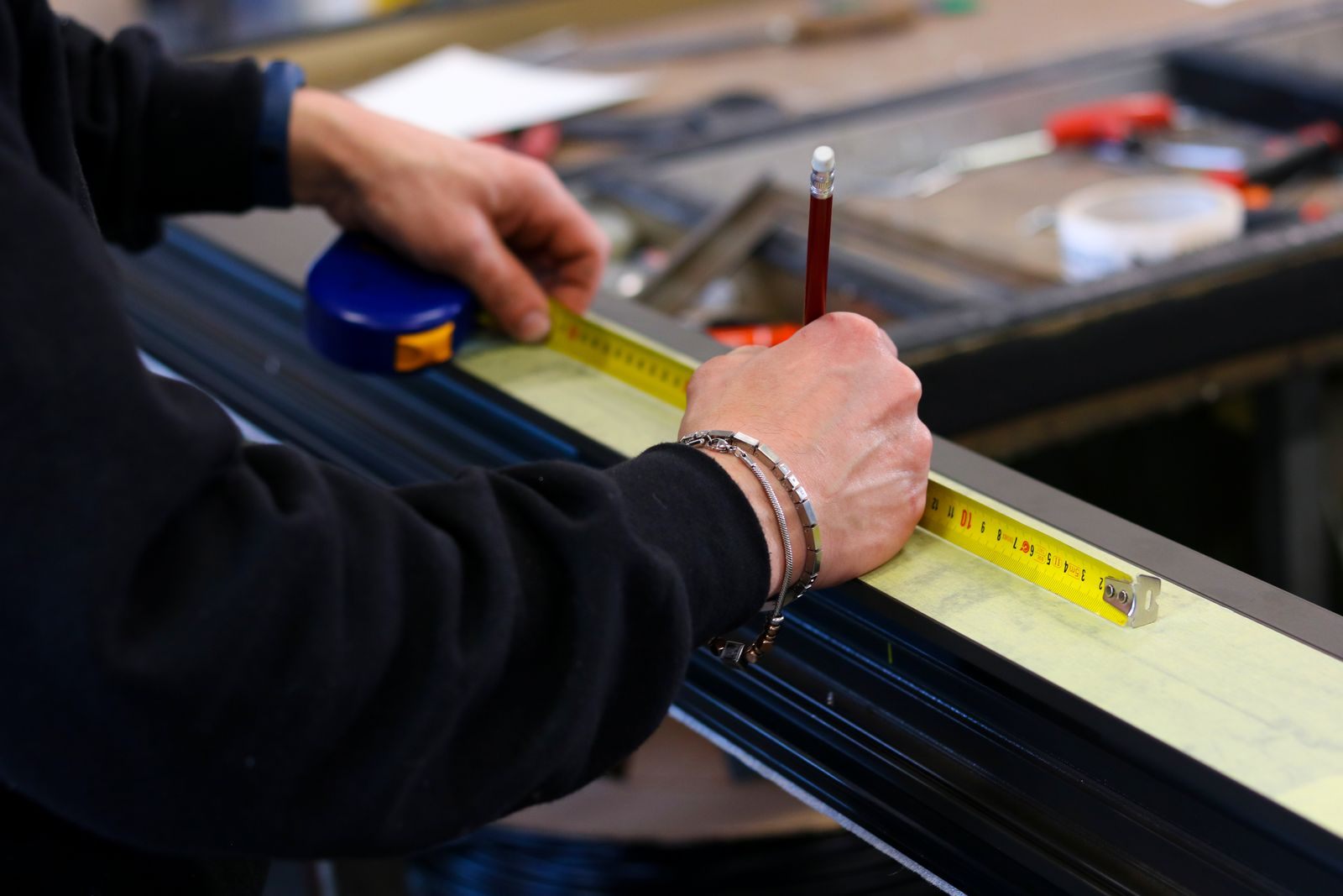 Worker measuring a dark aluminum frame section with a yellow tape measure and pencil in a workshop